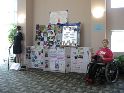 Large posters flanked by mannequins: one seated in a wheelchair, the other standing with the aid of a cane and with one arm missing.
